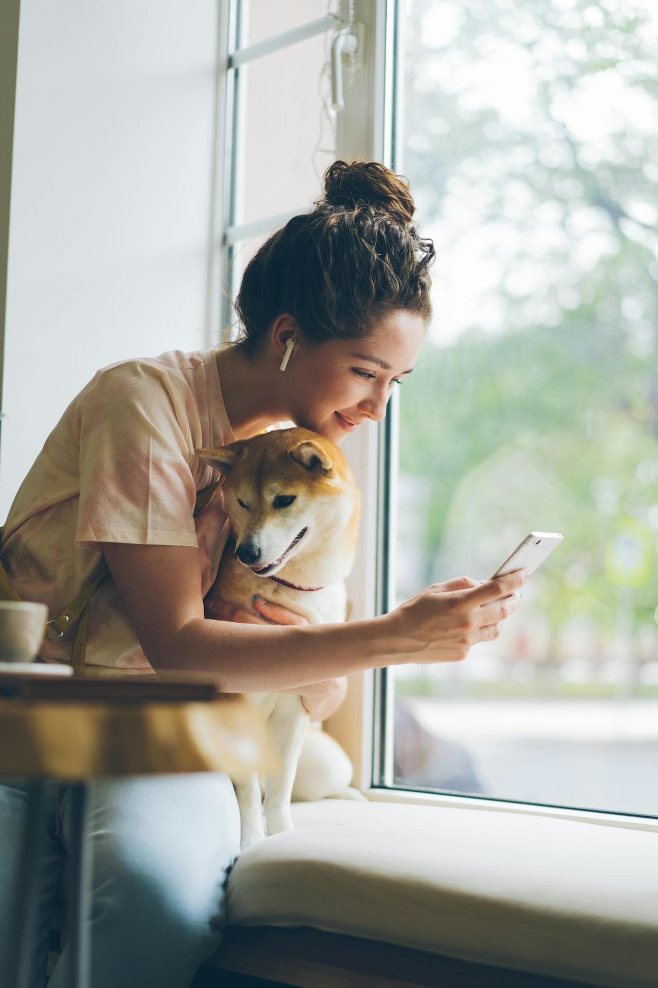 A dog owner scrolling through phone with dog on lap near window.